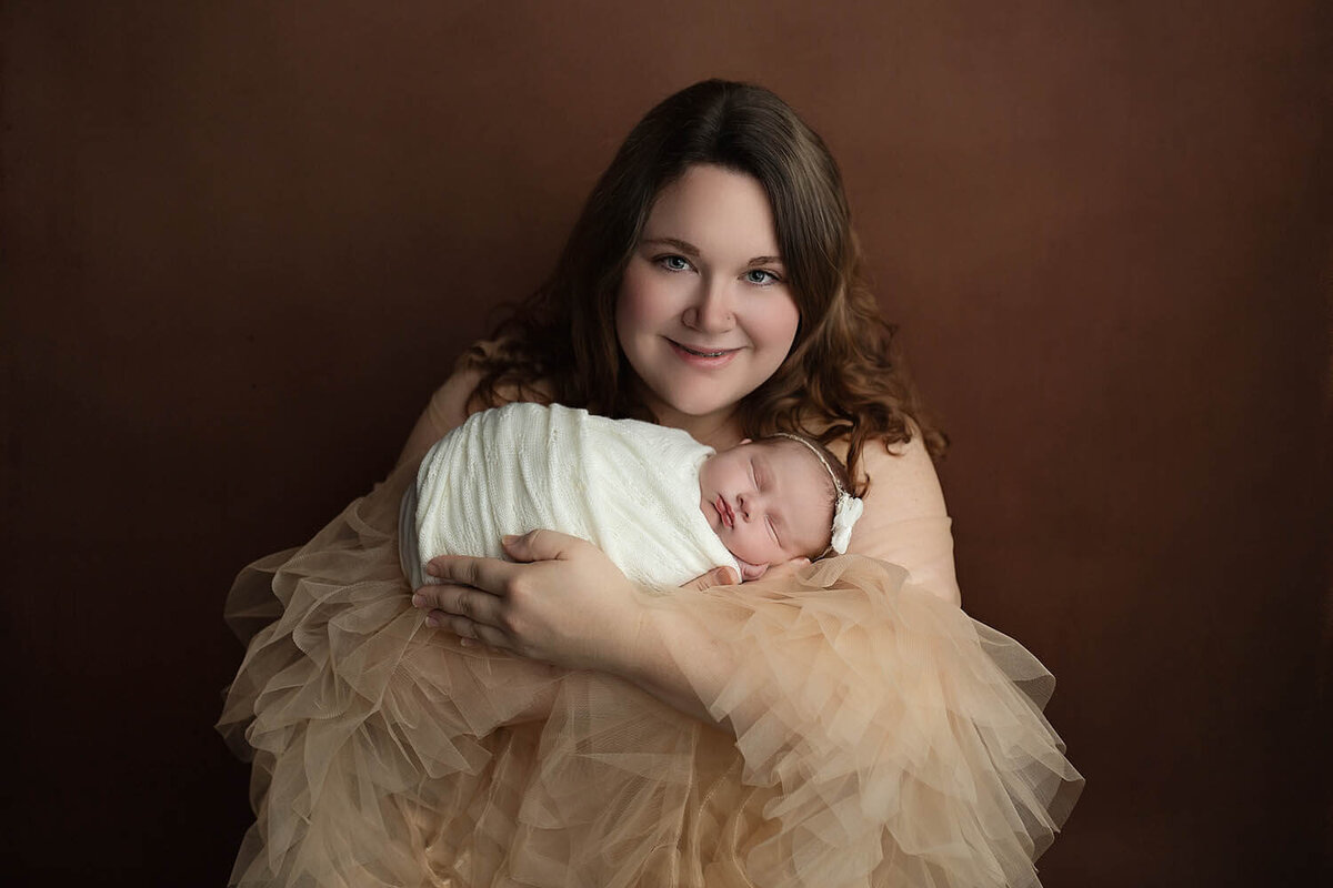 A happy new mother stands in a studio cradling her sleeping newborn baby in a tan tule dress