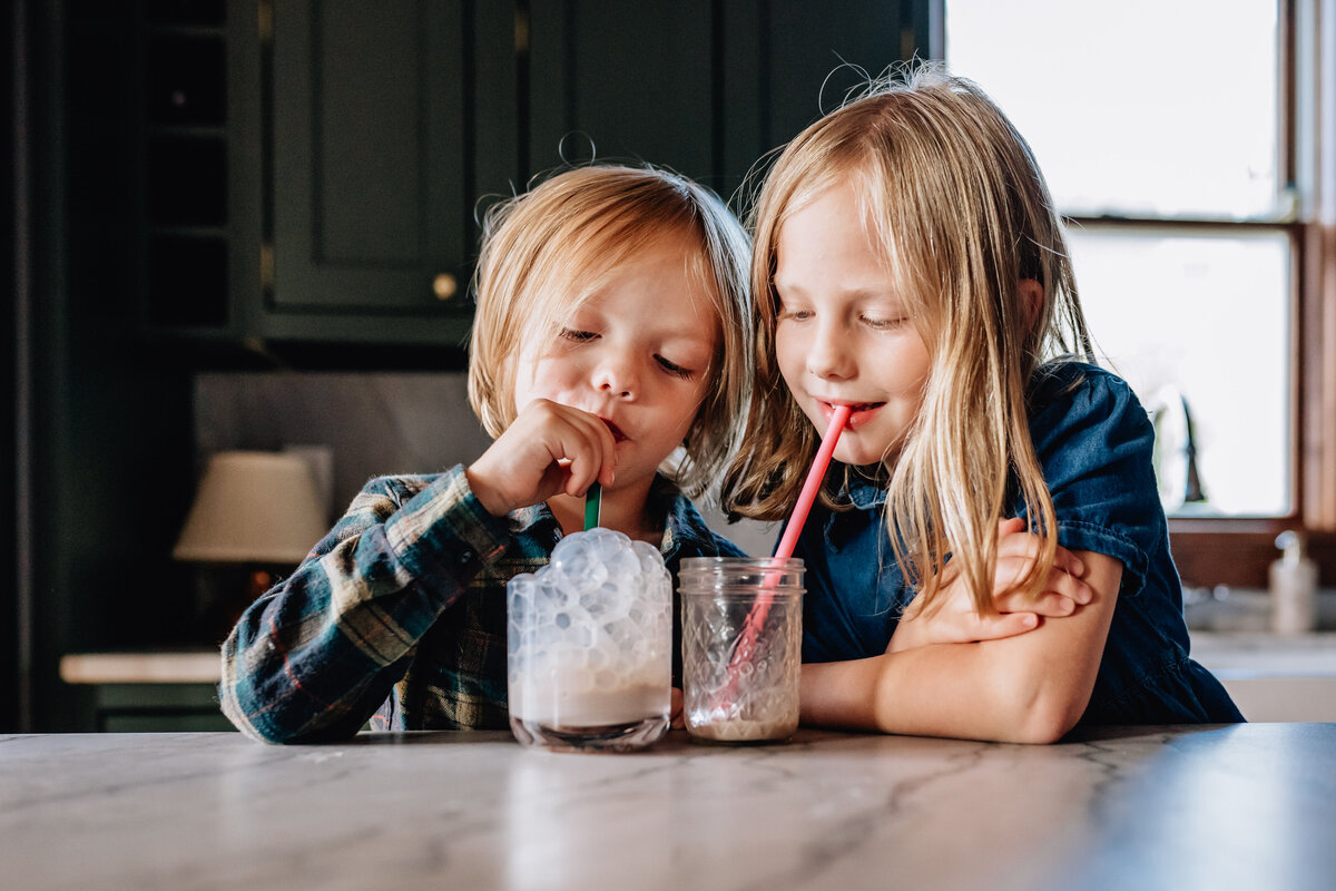 Two children sitting at a kitchen island, sipping drinks through colorful straws from glass jars, looking pleased as they share a moment together.