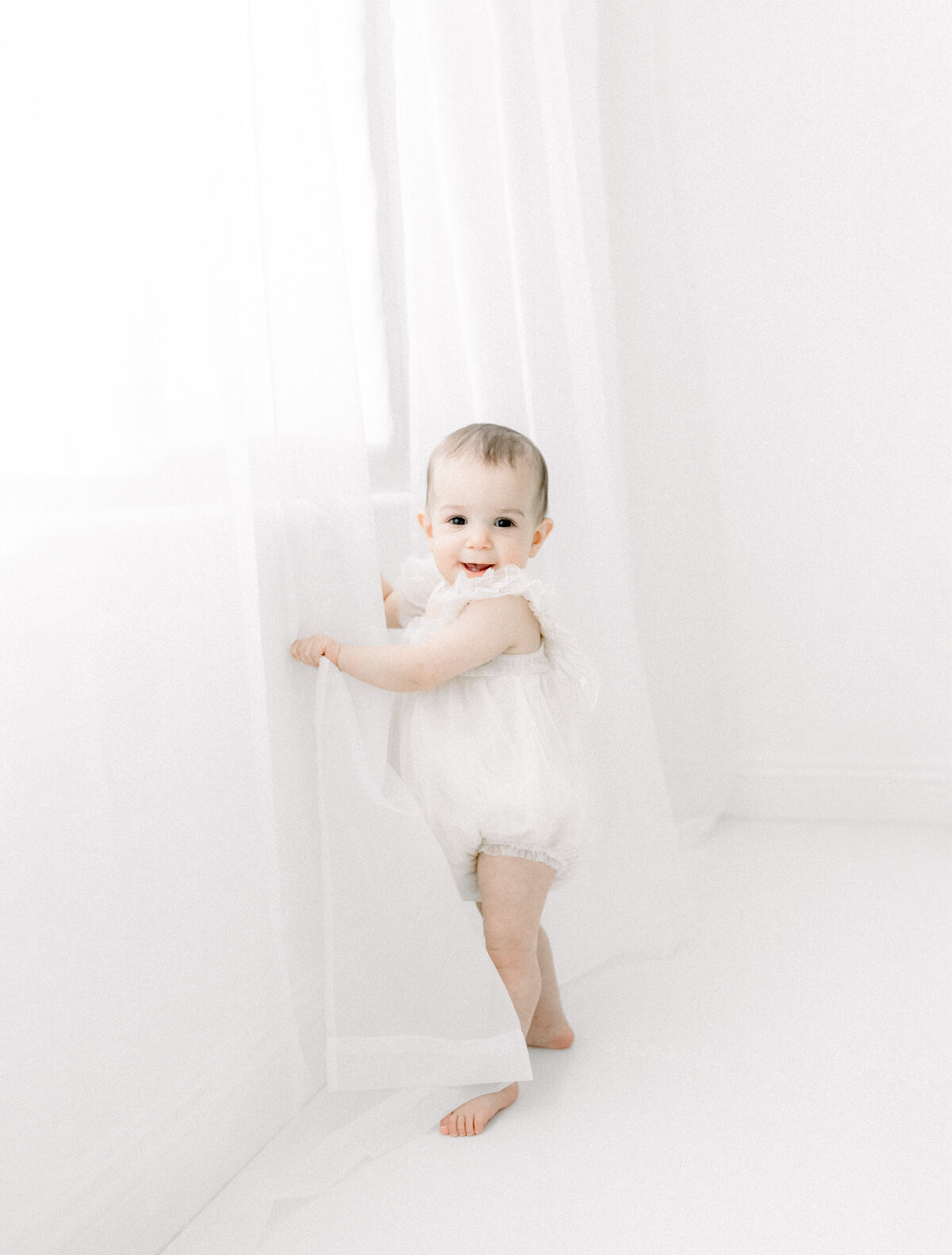 A baby girl standing up by a window holding the drapes