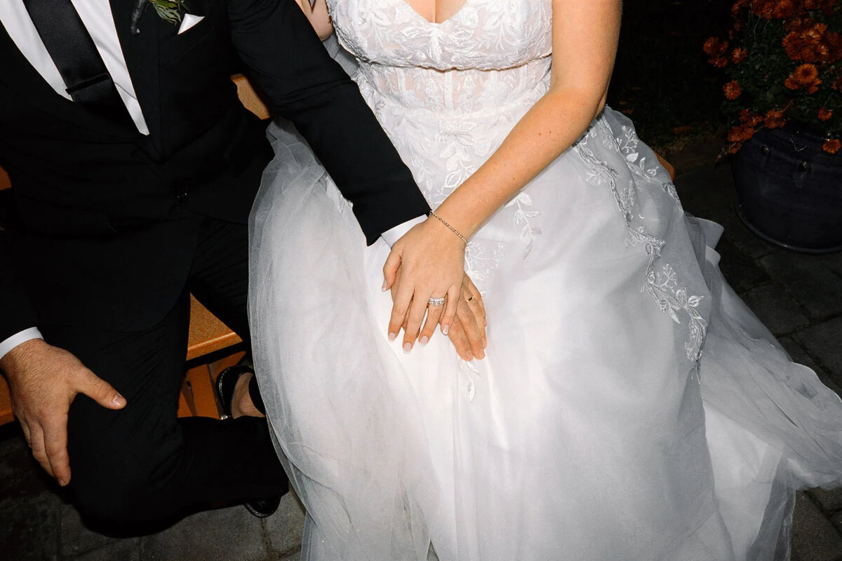 A bride in a white wedding dress and a groom in a black suit sit closely together, their rings visible, beautifully captured by a film photographer NJ—perfectly preserving this tender newlywed moment.