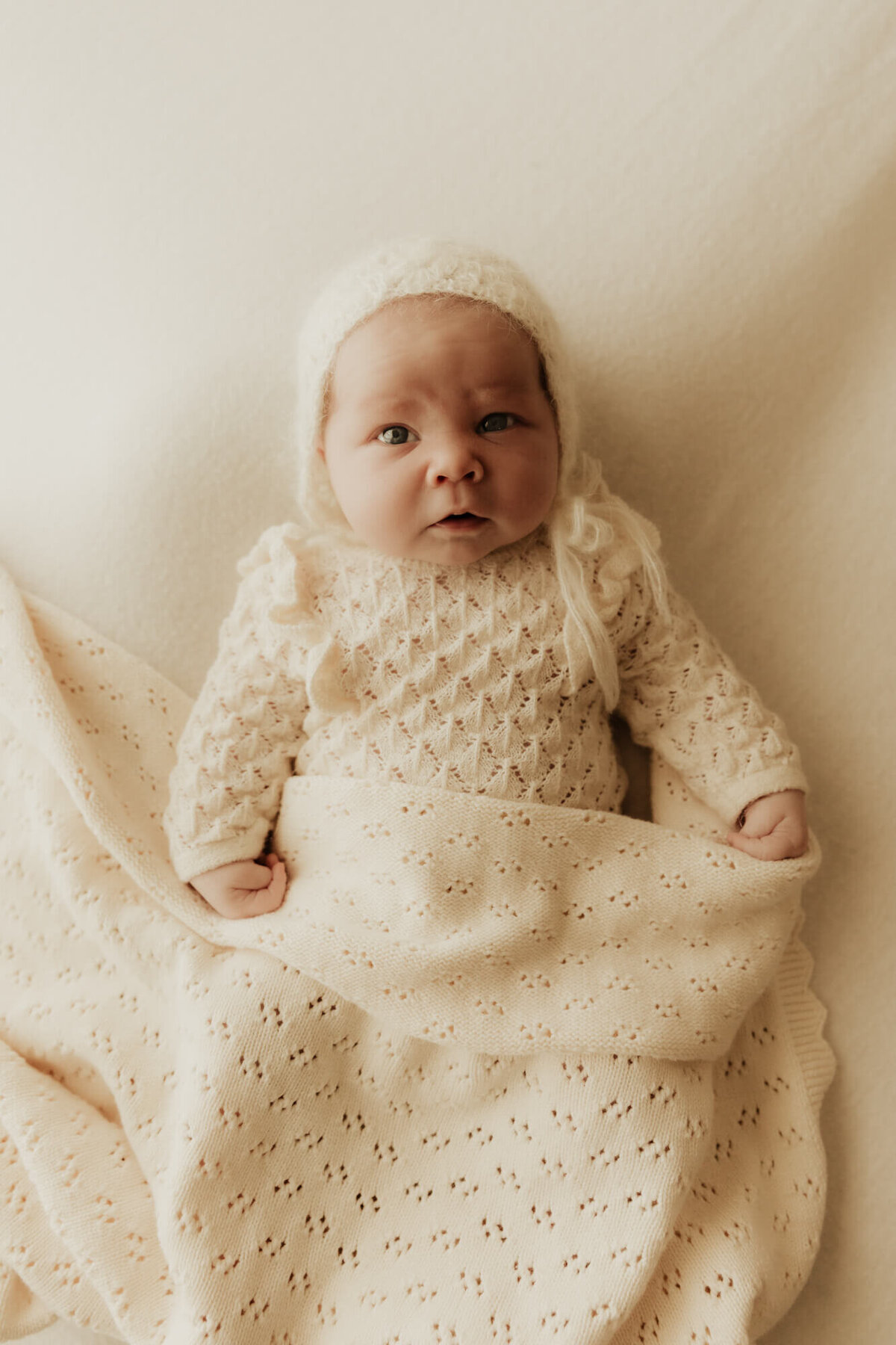 Newborn baby girl looks lies on a beanbag wearing a cream romper and bonnet while wrapped in  blanket.
