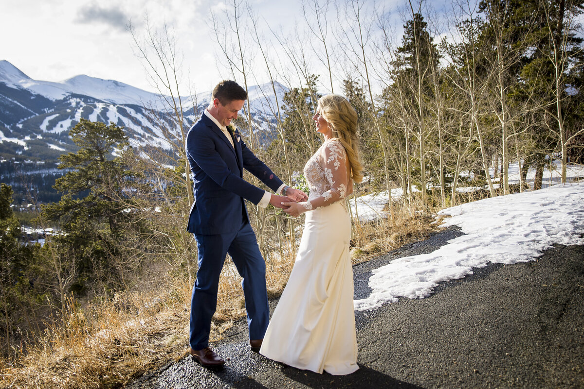 A bride and groom hold hands as the groom admires his bride's dress during the first look with a snowy mountain landscape in the background.