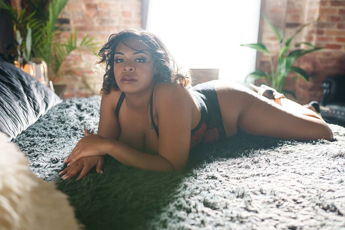 Dark skinned woman posing for a boudoir photoshoot wearing a red and black bodysuit posed laying on a bed with grey fuzzy sheets 