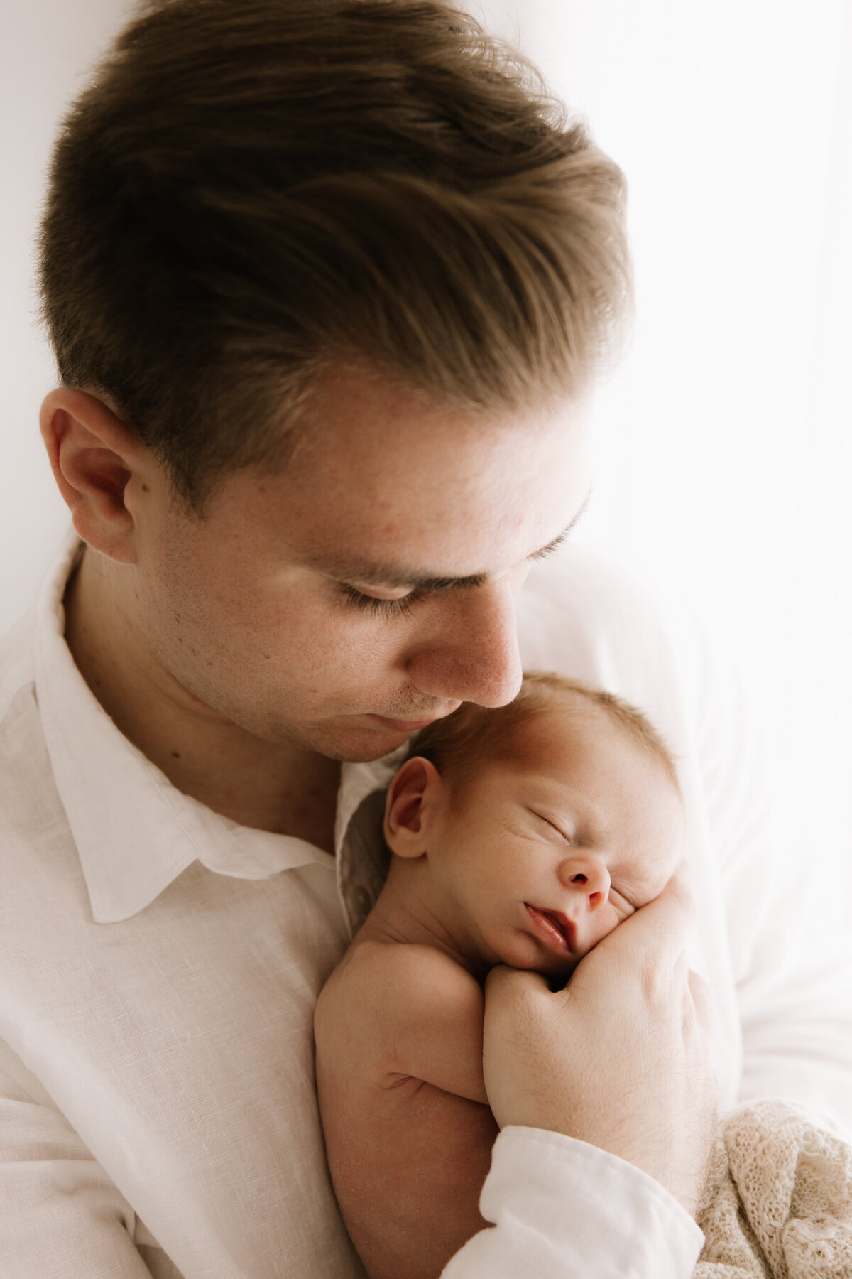 A close-up of a father gently cradling his newborn baby in his arms during a newborn photography session in Sydney. The father wears a white shirt and looks down at his sleeping baby with love and tenderness. The baby rests peacefully against his chest, wrapped in a soft textured blanket. The image is softly lit using diffused artificial light, creating a calm and intimate atmosphere.