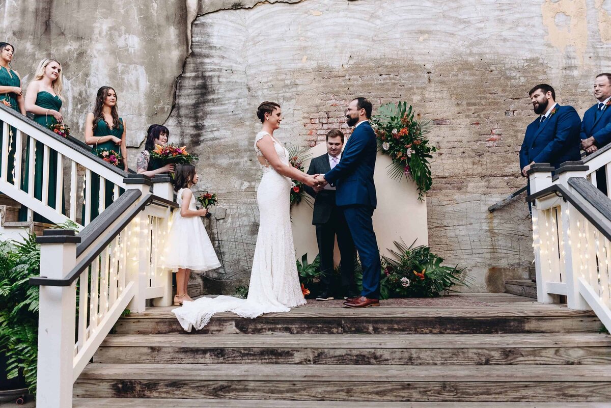 A bride and groom stand hand-in-hand at the center of an outdoor wedding ceremony, flanked by bridesmaids in green dresses and groomsmen in blue suits on the stairs, with an officiant and flower girl nearby.