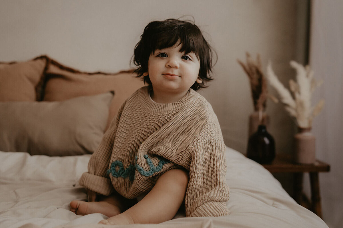 A 1 year old boy sitting on a bed in an oversized sweater in rochester ny