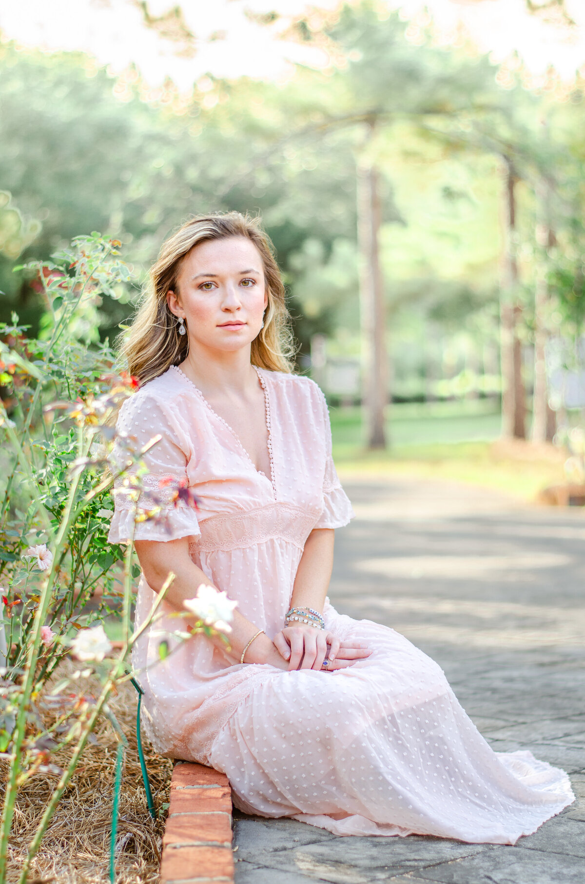 woman sits on the ground in a rose garden wearing a light pink dress