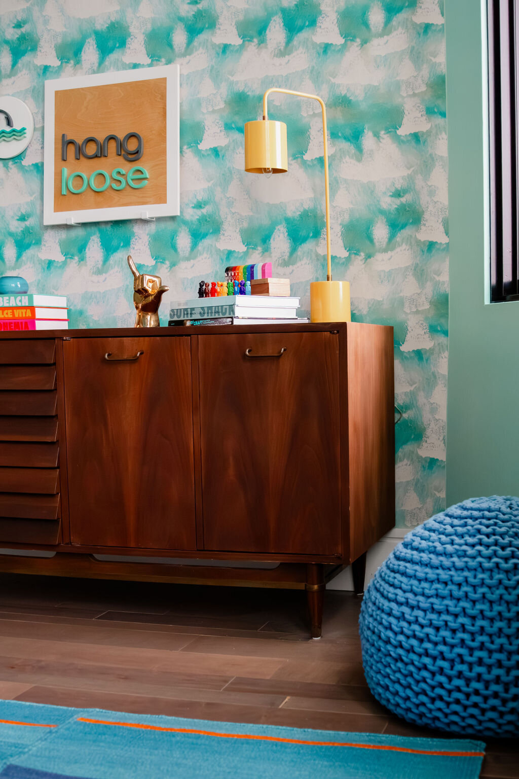 Mid-century modern wood dresser styled against a bold aqua and white abstract wallpaper in a surf-inspired kids’ room. Decor elements include a yellow modern desk lamp, colorful books like Surf Shack and Capri Dolce Vita, a shiny gold shaka hand sculpture, and a playful wall sign that reads “hang loose.” A bright blue knit pouf and turquoise rug add cozy texture and vibrant color, perfect for a beach-themed bedroom with a modern maximalist twist.