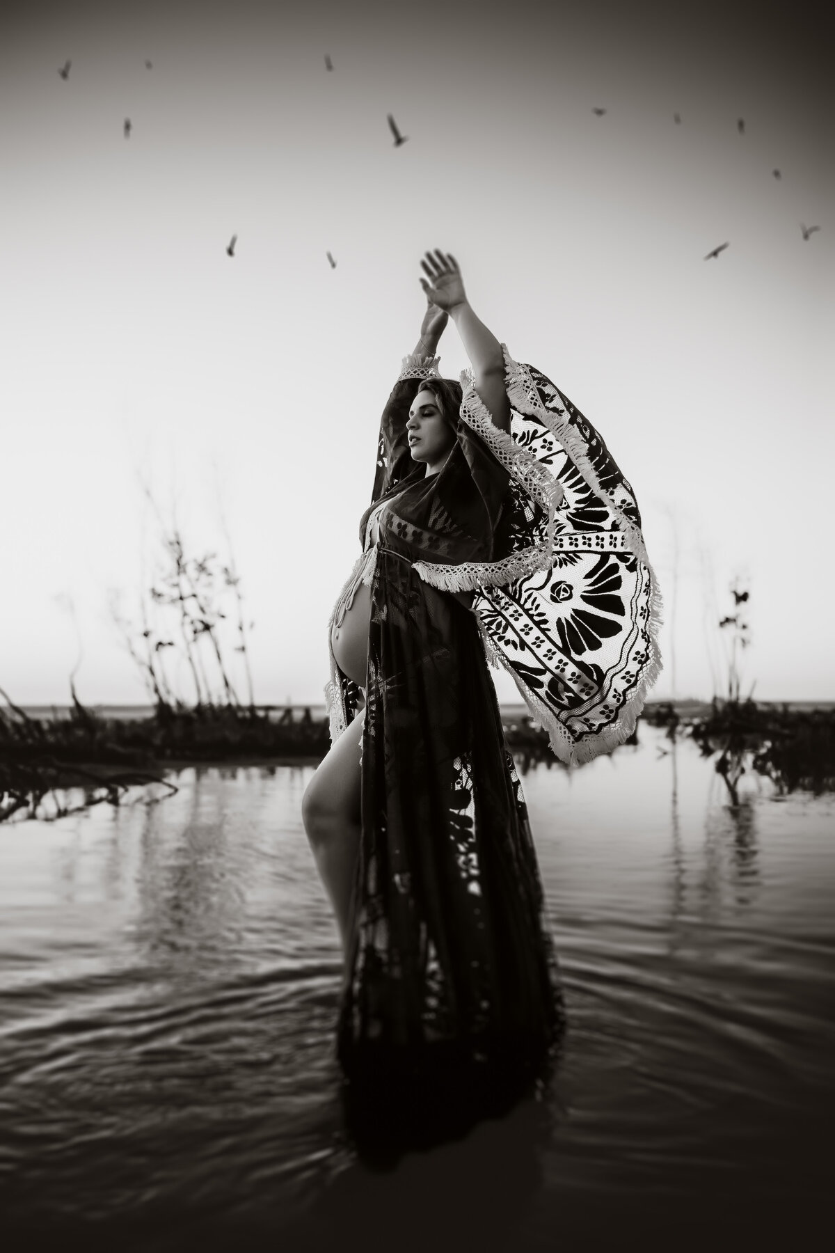 black and white maternity portrait of a women in a bpho crochet dress standing in the water with birds flying above