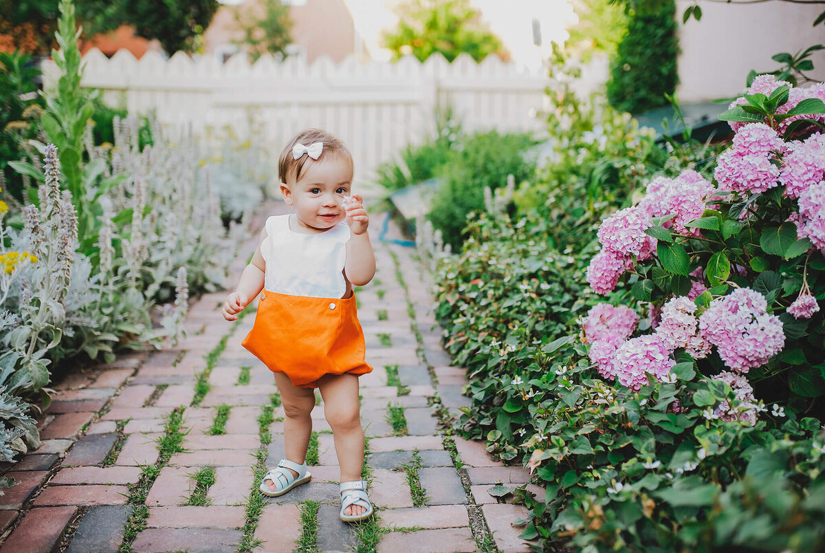 Baby girl in orange shorts smelling a flower on a brick path in Fells Point Baltimore Maryland