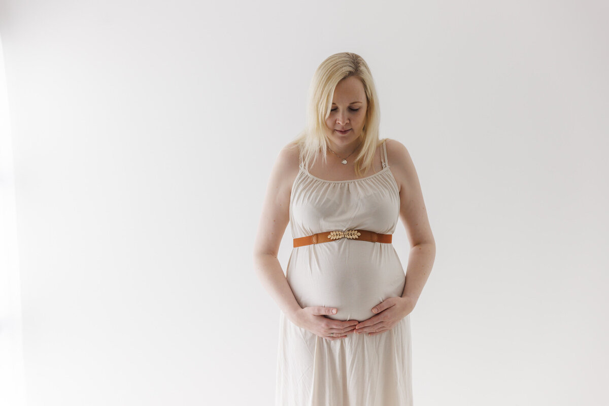 En gravid kvinne står vendt mot kamera og omfavner magen sin. Iført hvit kjole og brunt belte over magen. Fotografert i Studio Landmark, Fyllingsdalen.