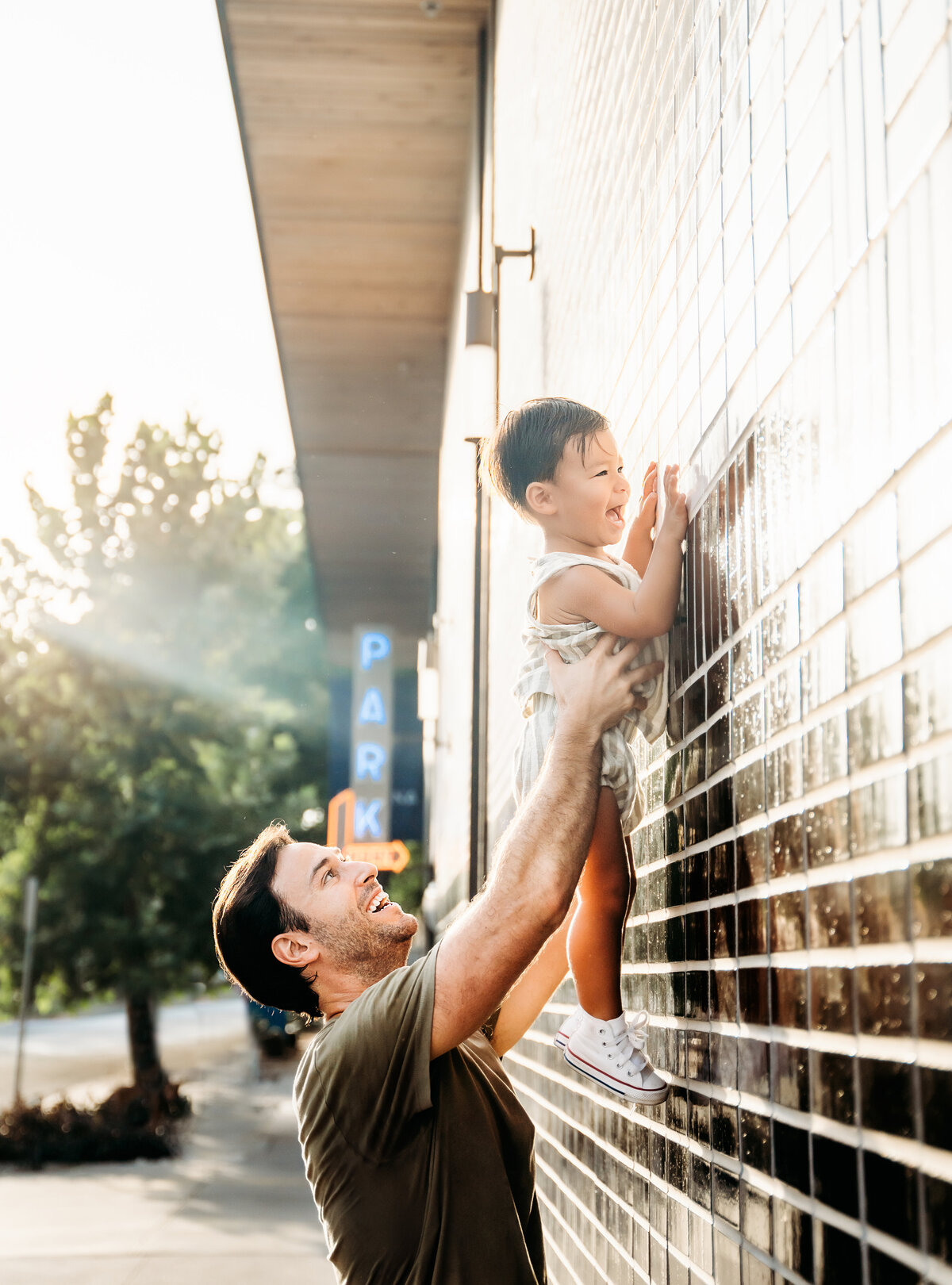 Family Photographer, dad lifts baby boy up to examine a tiled wall outside, both laugh