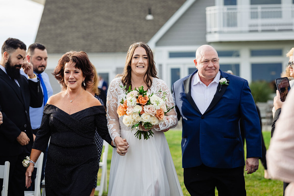 A bride being escorted down the aisle by her parents; her father in a blue suit and her mother in a black dress. She holds a bouquet of orange and white flowers, smiling as guests watch.