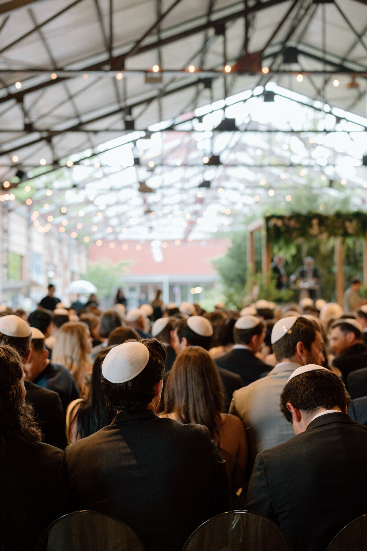 Crowd view of Jewish wedding ceremony under a glass venue with greenery.
