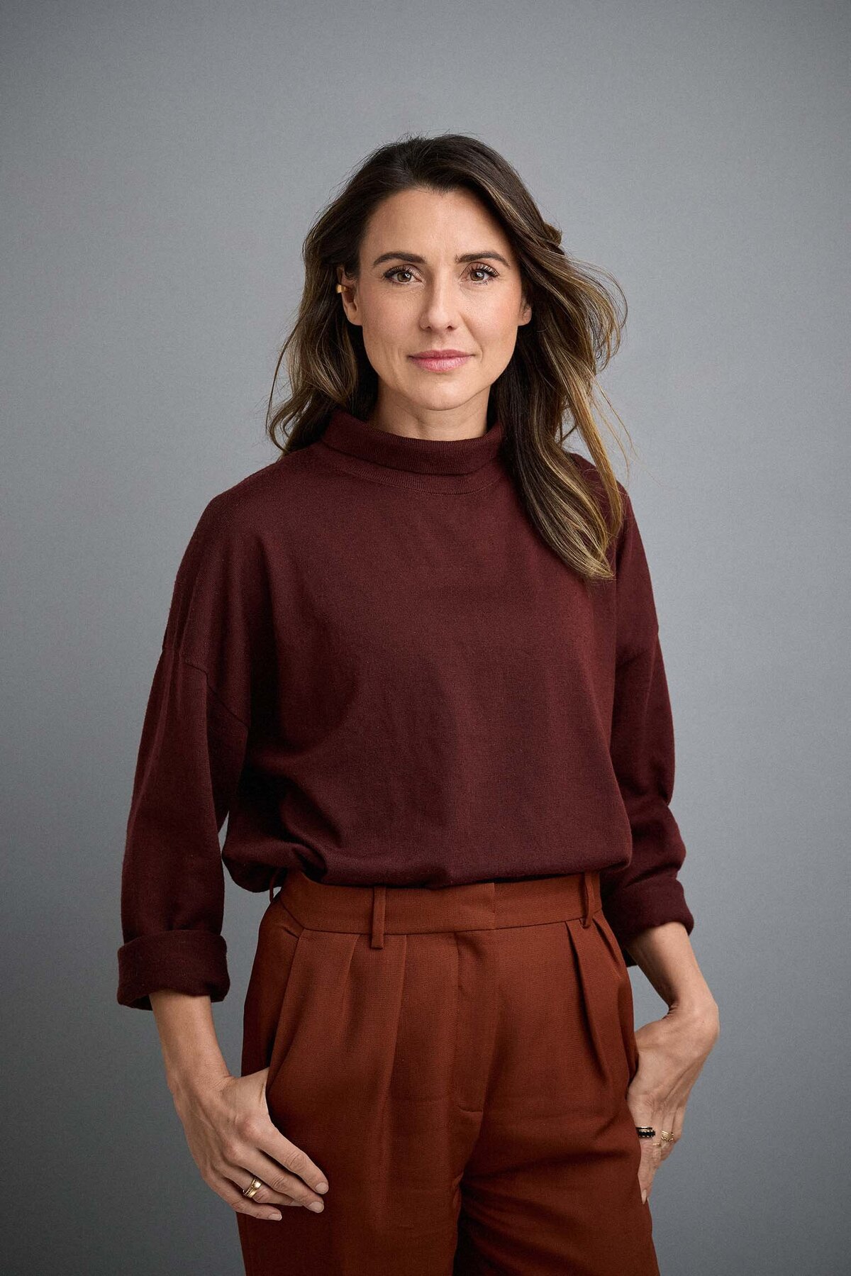 Confident woman with hands on hips wearing burgundy jumper and rust trousers. Against a grey studio background.