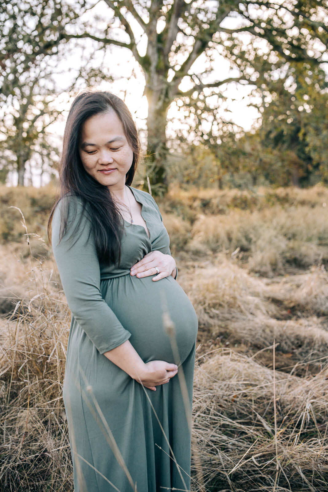 Mom to be standing in a field wearing a green maternity dress and looking down her shoulder, hands snuggled around her belly