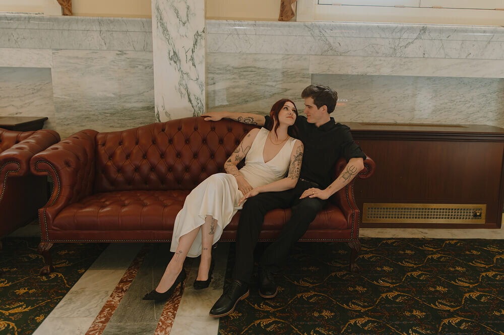 Sitting on a red leather couch inside of a government building, a man and woman lovingly look at each other.