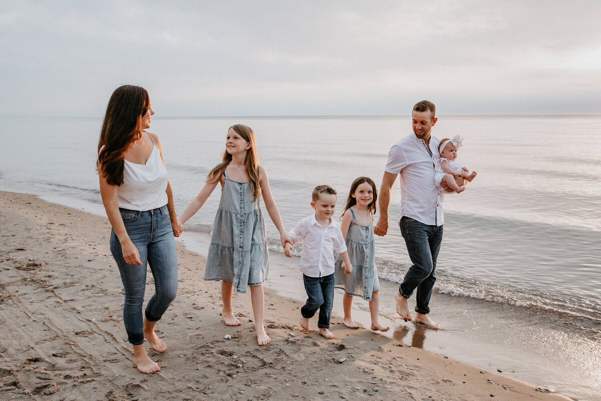 Mom, Dad, and four children are walking hand-in-hand for a top family photoshoot at Grand Bend beach. The family is holding hands, with mom and dad on the outside and three young children between them. Dad is holding the youngest. They are walking in a line and laughing.
