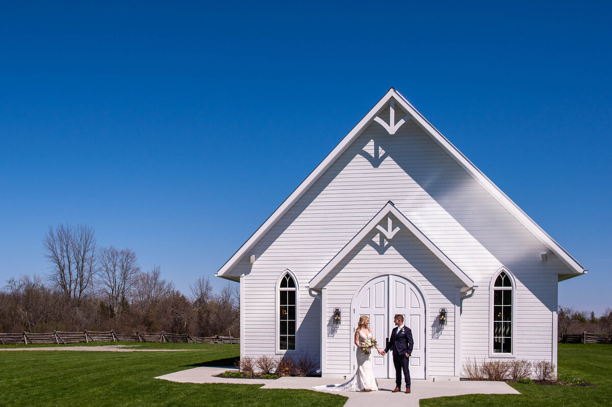 a scenic photo of Stonefields Estate Ceremony House showing  big blue sky and and an elegant bride and groom outside the front doors.  Captured by Ottawa wedding photographer JEMMAN Photography