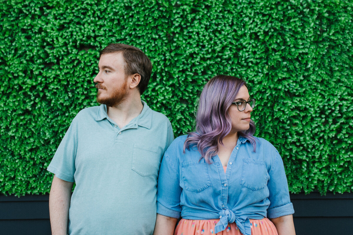 A couple standing next to each other while looking in opposite direction in front of a large hedge during their engagement session in Fort Worth, Texas. The woman on the right has silvery purple hair and is wearing a jean jacket, a salmon skirt with colorful polka dots, and glasses. The man on the left is wearing a light blue, short sleeve, collared shirt.