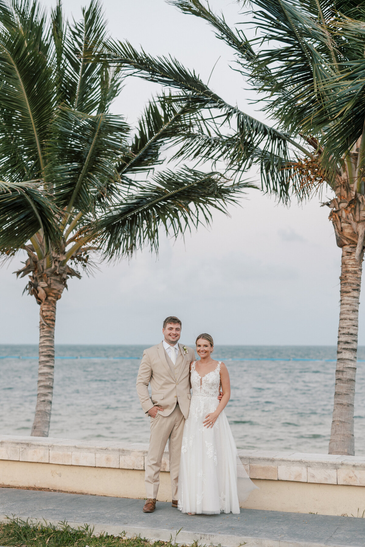 Bride and groom pose seaside at their Cancun destination wedding, palm trees swaying behind them.