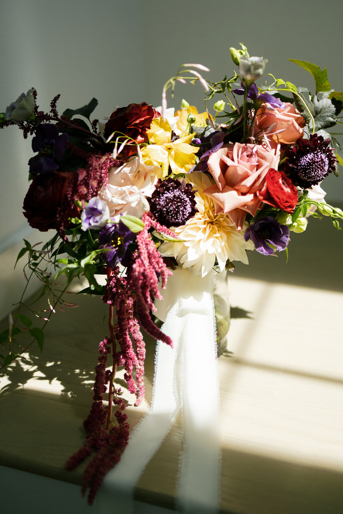 Vibrant bridal bouquet featuring burgundy ranunculus, yellow poppies, peach roses, purple lisianthus, and trailing raspberry amaranthus, photographed in dramatic natural light for artistic wedding detail inspiration.