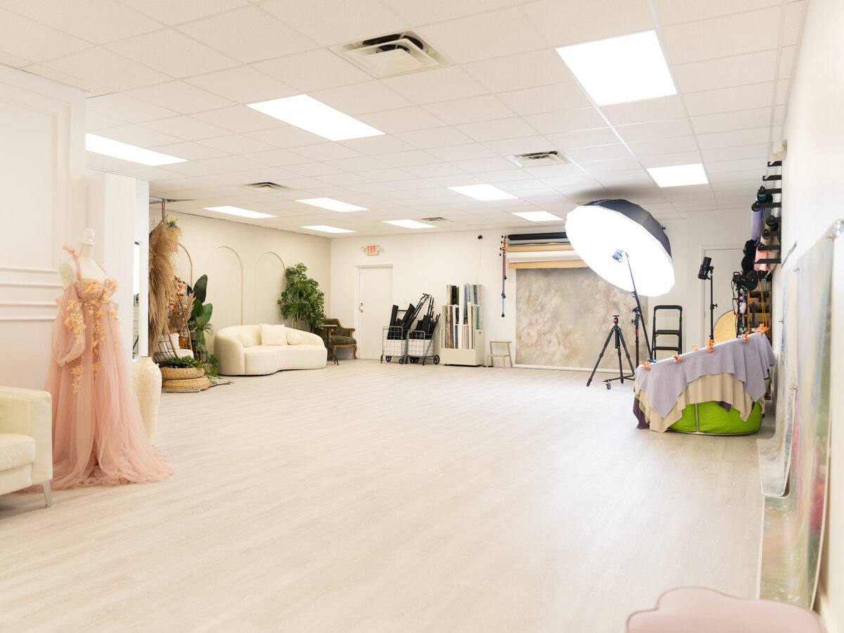 A spacious, well-lit photography studio features props, a white backdrop, and a mannequin in a pink dress. The atmosphere is creative and professional.