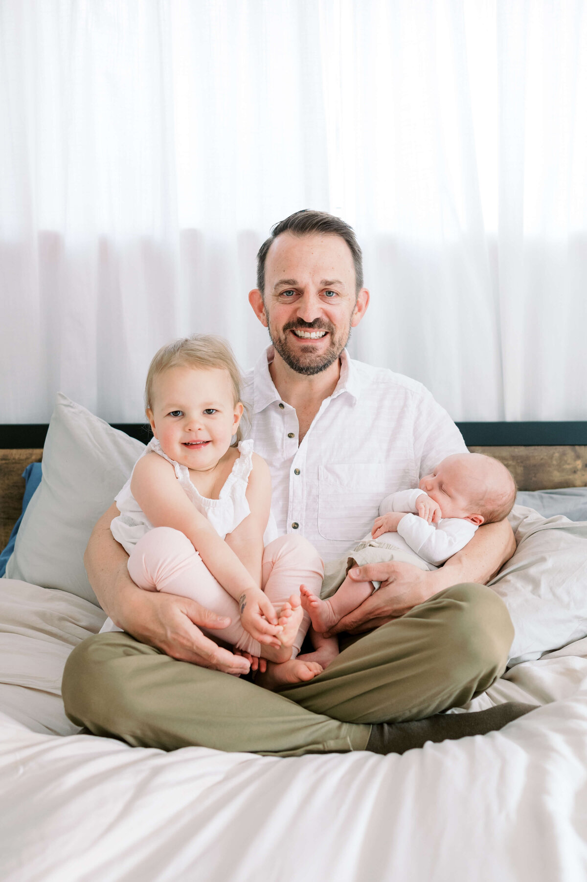 A well dressed man holds his young daughter and new son while sitting on his bed