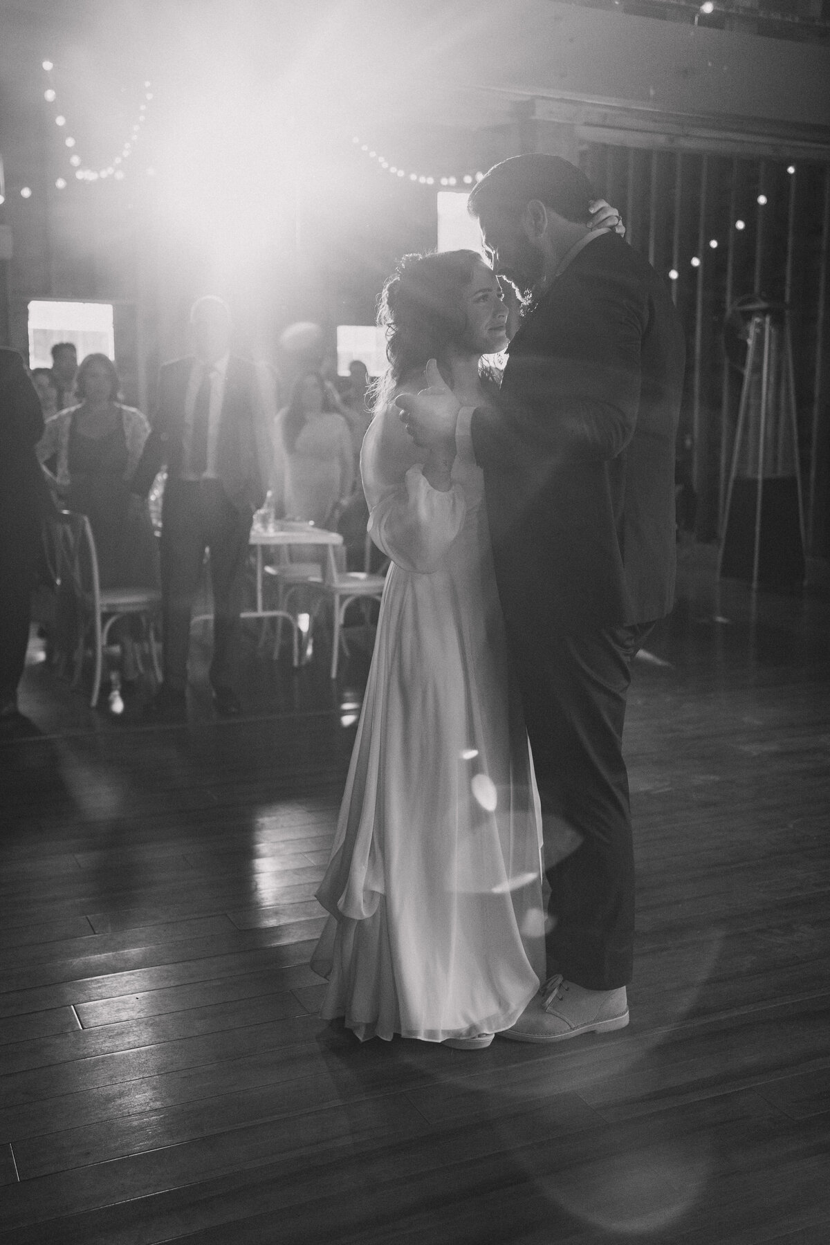 Bride and groom share a tender first dance in a beautifully lit venue, with soft string lights creating a romantic ambiance. The bride, in a flowing gown, and the groom, in a dark suit, are captured in a heartfelt, intimate moment, surrounded by guests who look on with admiration. This black and white photo emphasizes the timeless and emotional essence of their special day.