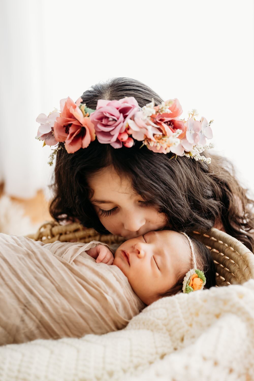 studio photography session with newborn baby being kissed by big sister. sister wears a flower crown