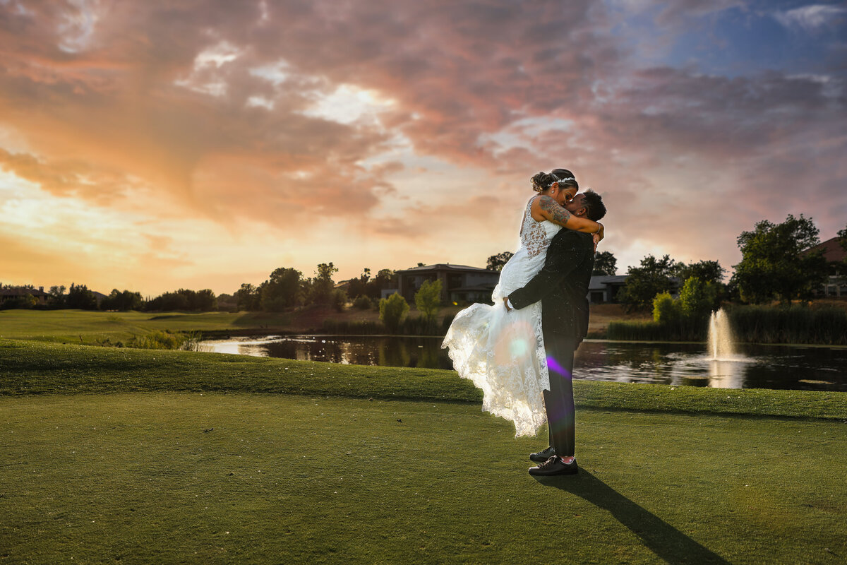 Groom lifts bride up on golf course in front of pond with cloudy sky in the background. Photo by Sacramento wedding photographer, philippe studio pro.