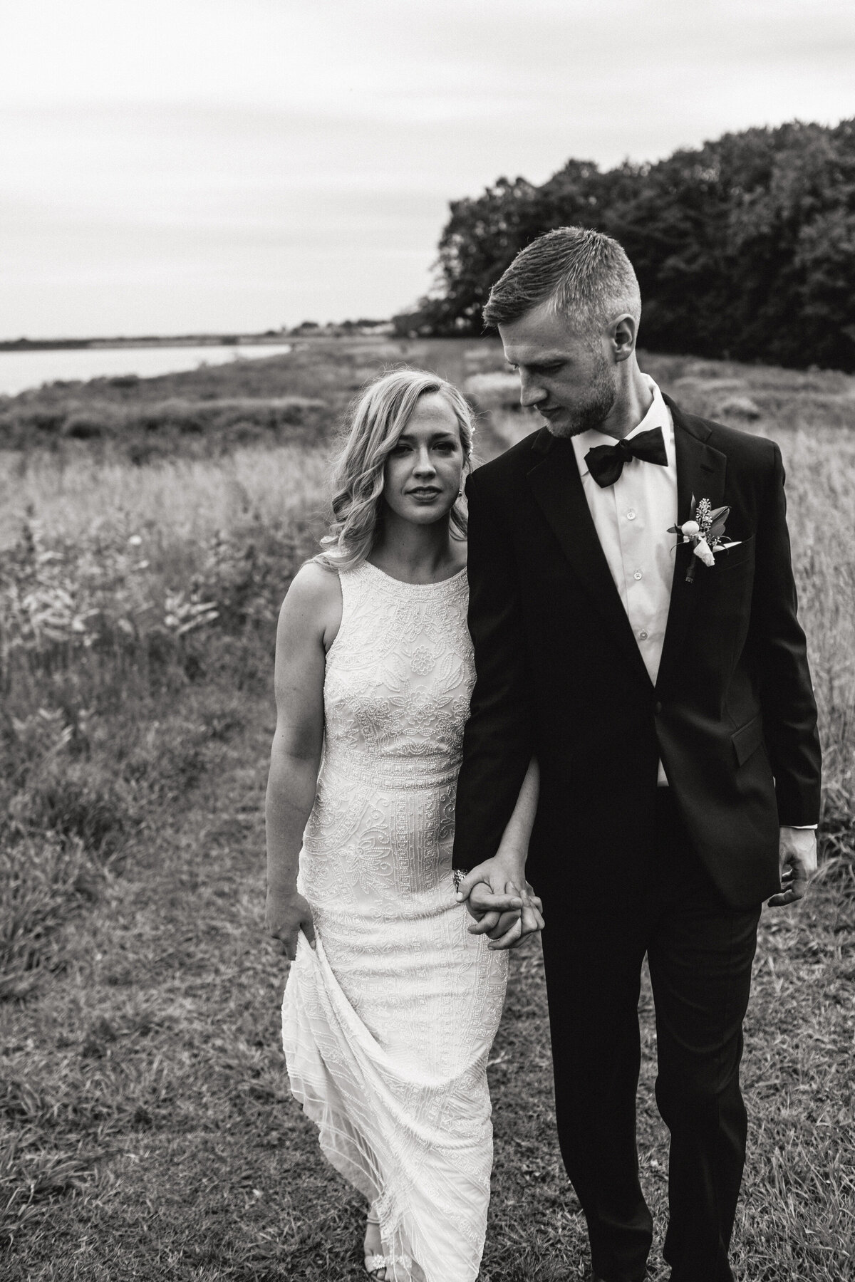 A black and white photograph of Lindsay and Tom on their wedding day at Thompson Island in Boston, Massachusetts. The bride and groom are walking towards the camera, hand-in-hand, through a field of natural grass. The bride is looking into the camera and the groom is looking towards her. The groom is wearing a black tuxedo and the bride is wearing a beaded wedding dress. In the background is a stand of trees and the water of the channel. Wedding photography by Stacie McChesney/Vitae Weddings.