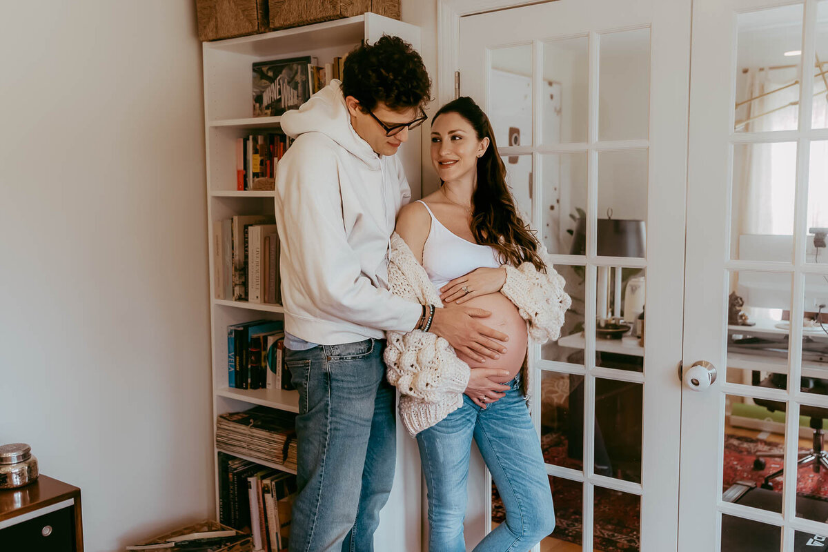 A couple stands in a room by a glass door. The person on the left, wearing glasses and a hoodie, is embracing the pregnant person on the right, who is wearing a cardigan. They look at each other.