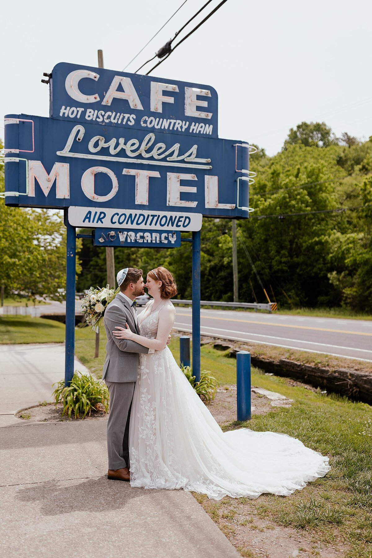 Redheaded bride wearing a flowing v-neck wedding dress with floral lace kisses the groom wearing a light gray suit  and white kippot in front of the Loveless Cafe sign at their Jewish wedding in Nashville, TN