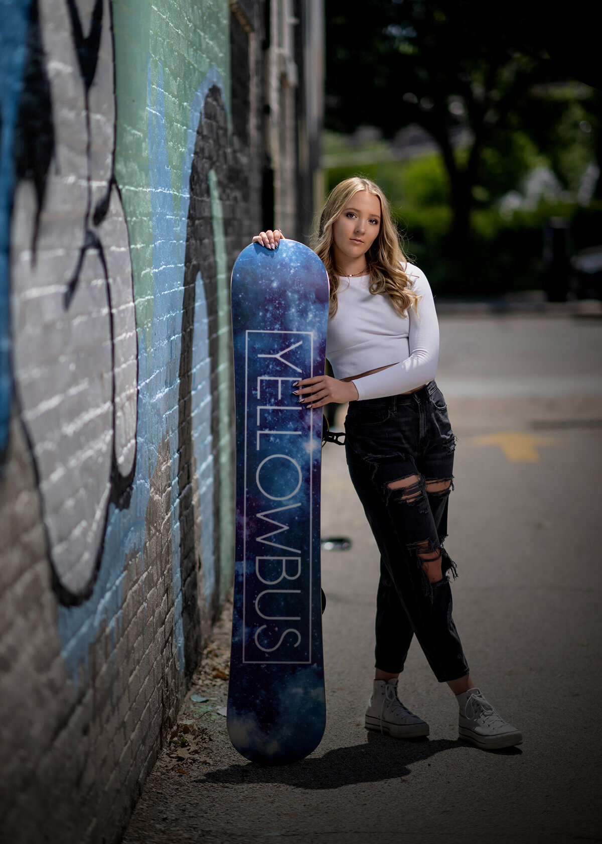 A high school senior leans on a snowboard in an alley in a white top and ripped jeans