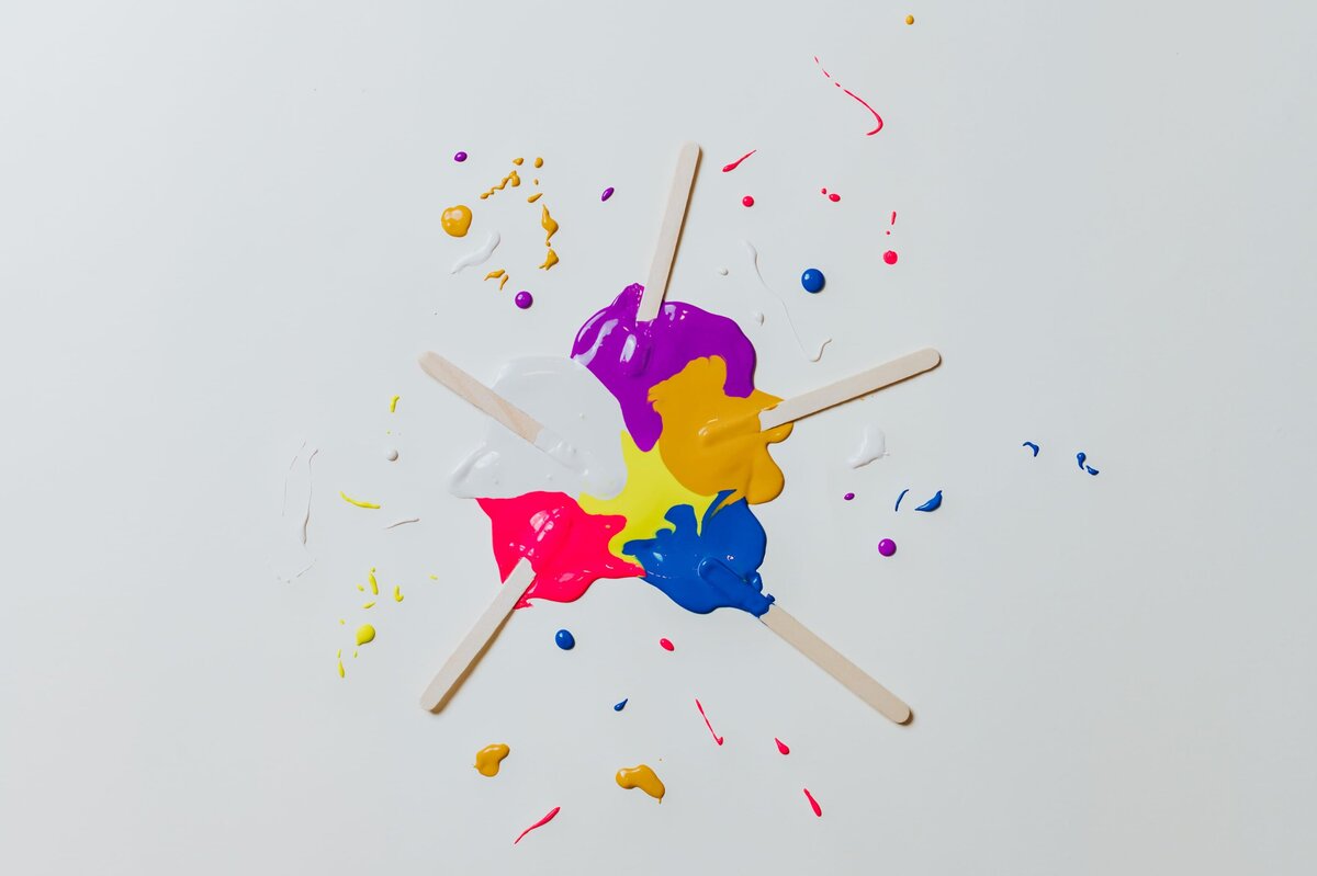 White backdrop with white, mustard, purple, yellow, pink and electric blue paint splattered in the middle with popsicle sticks in each color.