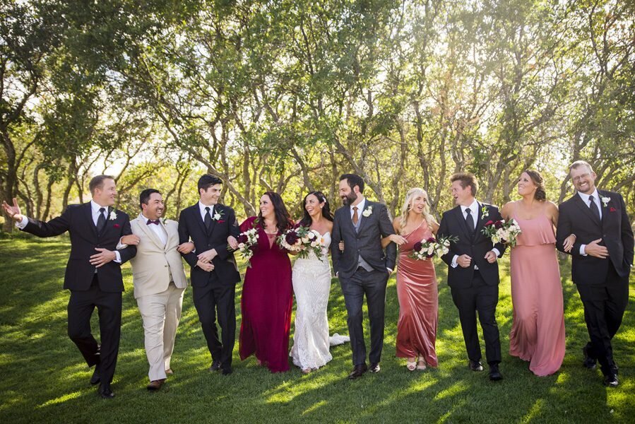 A bride and groom walk arm in arm with a long line of their wedding party as they all laugh with one another.