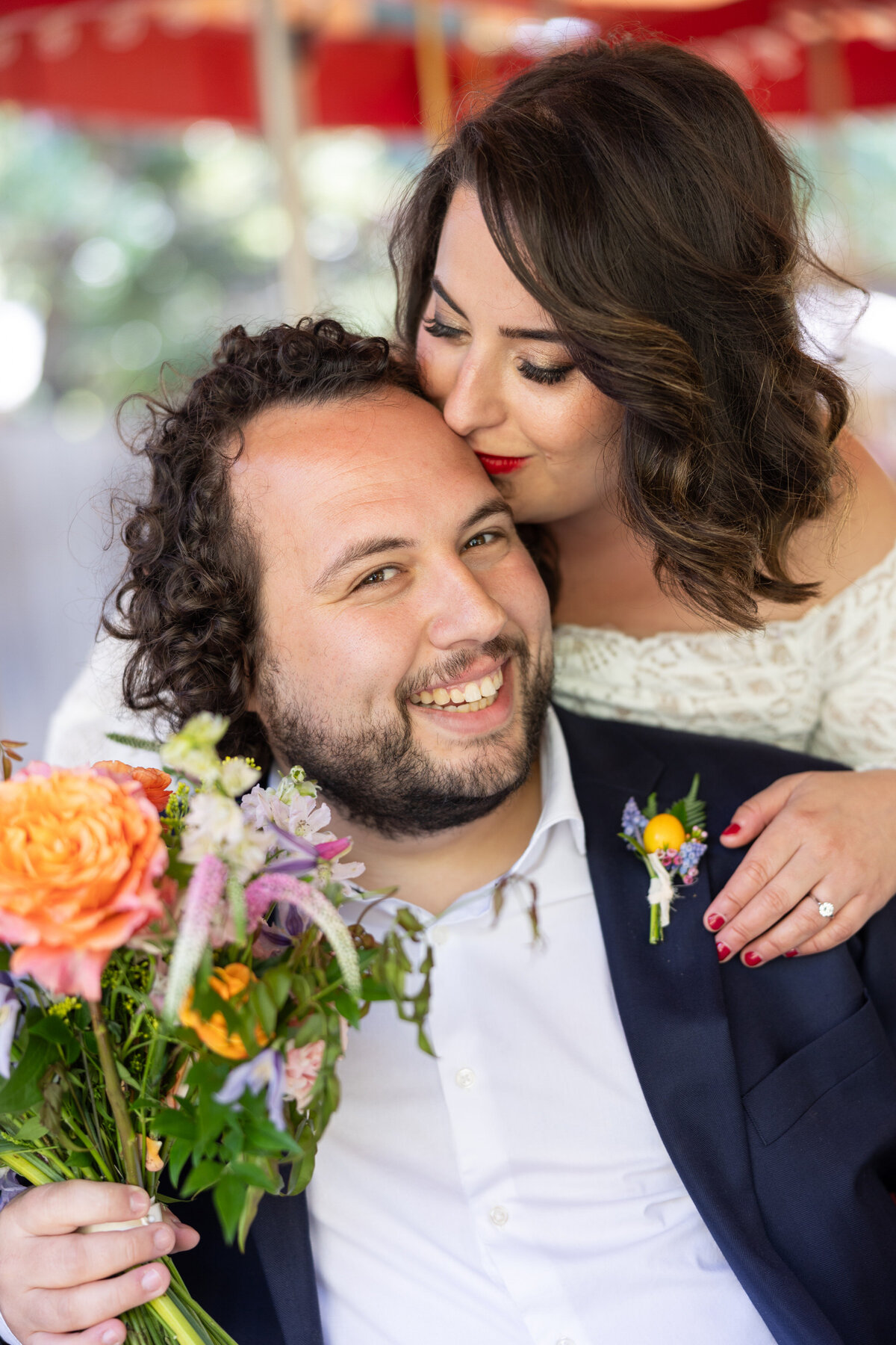 A bride standing behind a groom kissing their forehead