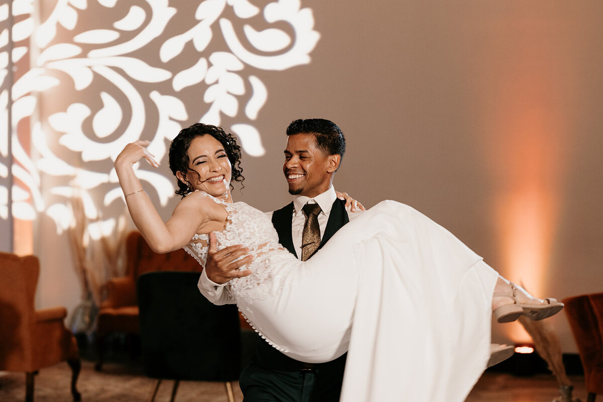 Groom joyfully carrying the bride on the dance floor with a romantic light pattern projected on the wall, highlighting their happiness.