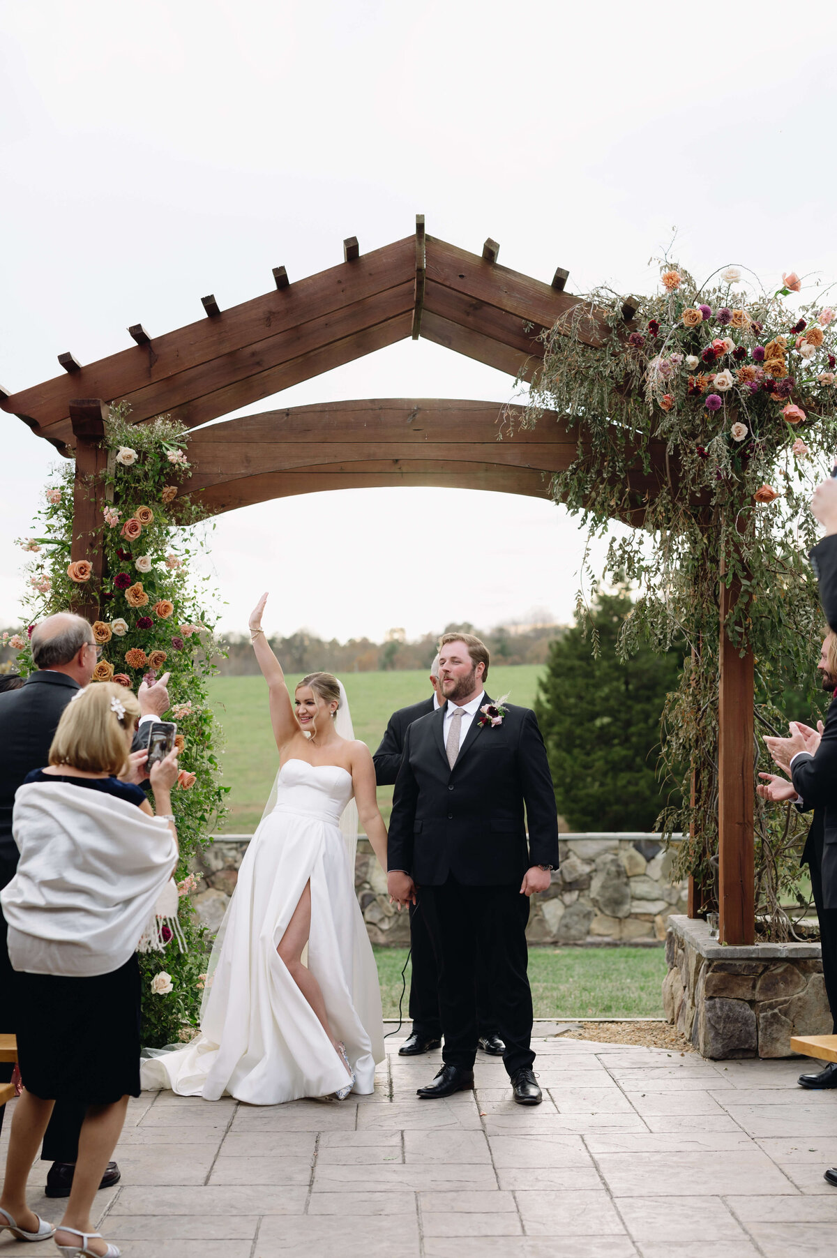 bride holding her grooms hand by her side while raising her other hand in the air in celebration with the groom smiling and grandparents taking pictures of the outdoor wedding ceremony with green hills in the distance and florals decorating the space