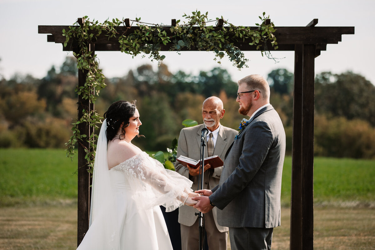 A bride and groom hold hands during their outdoor ceremony under wooden arbor with officiant