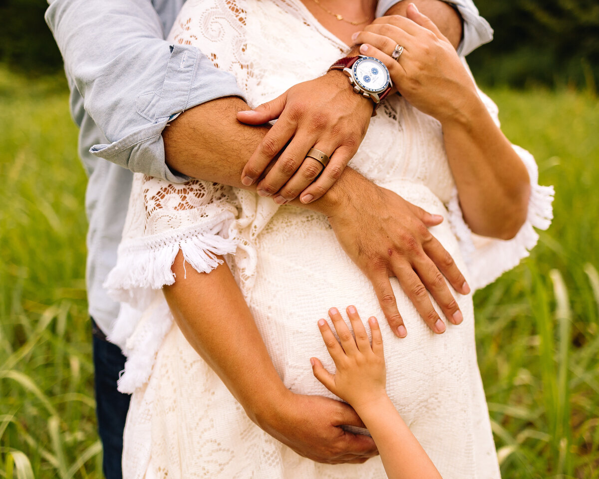 Photograph of a pregnant woman's belly with her family's hands. The man has a blue shirt and watch, the woman a white dress.