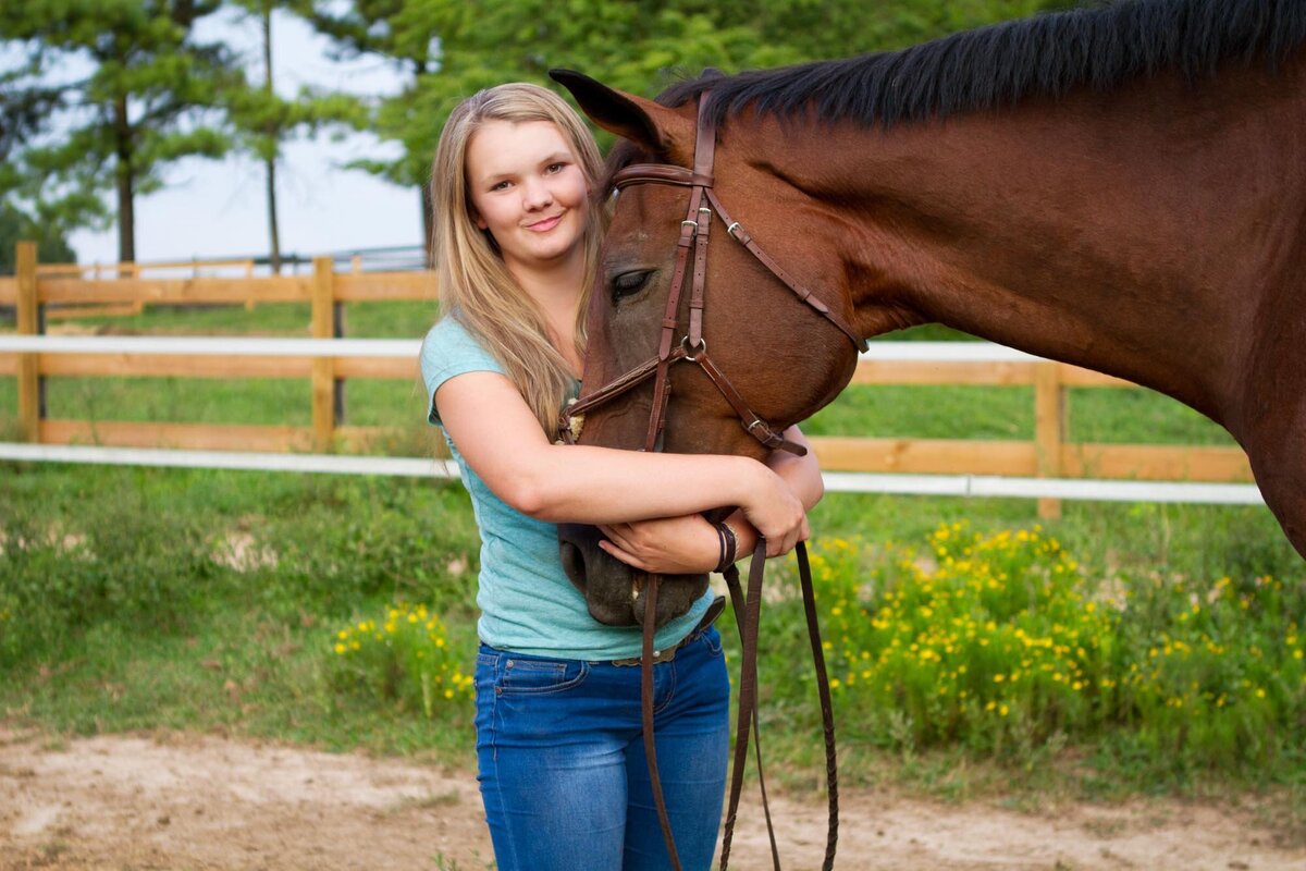 Senior portrait of a girl in a blue shirt hugging her horse with grass and flowers behind them.