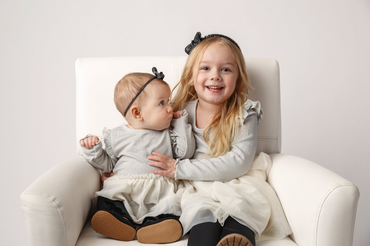 Image of a three year old girl sitting on a chair with her baby sister.