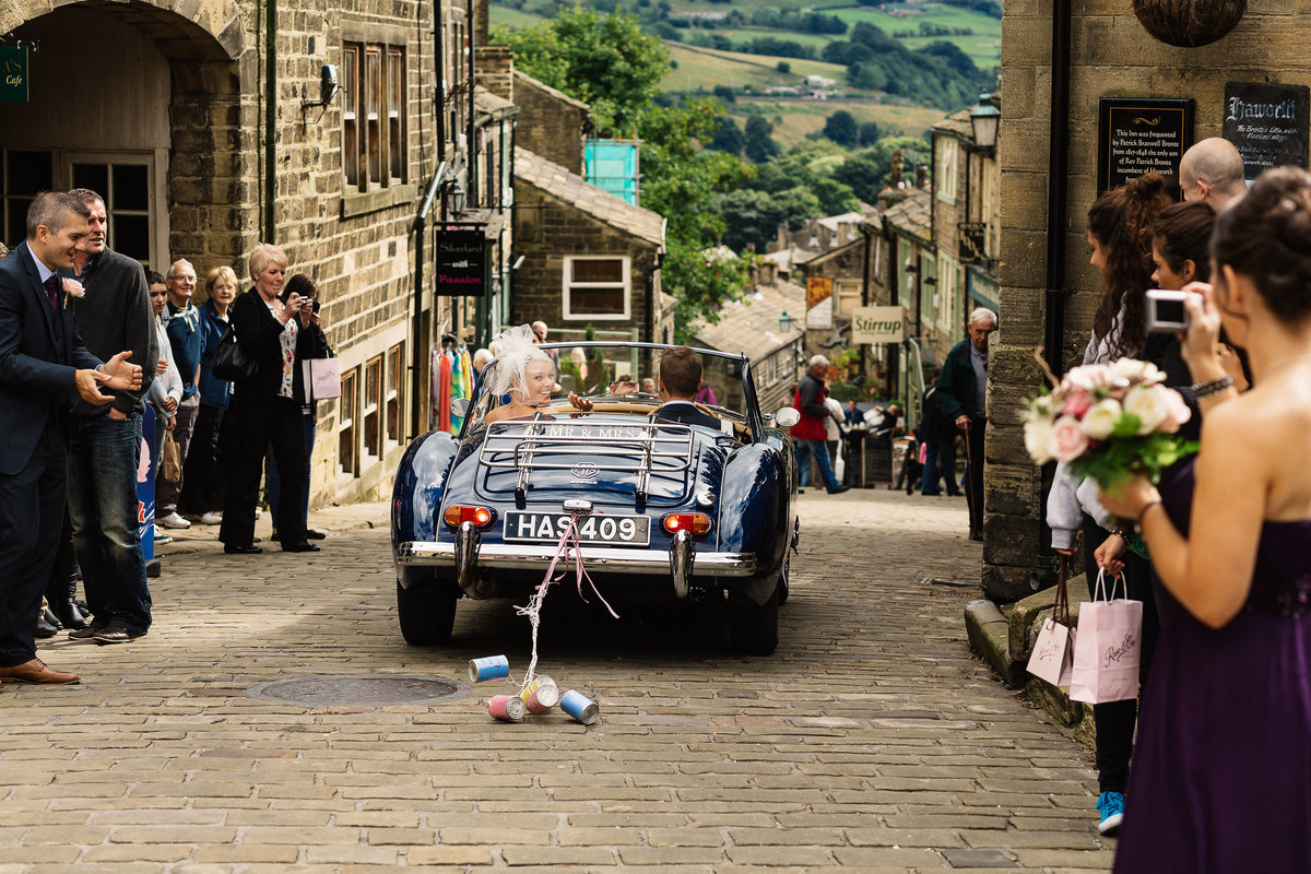 Bride waving at her guests as she leaves in a convertible wedding car on Haworth Main Street