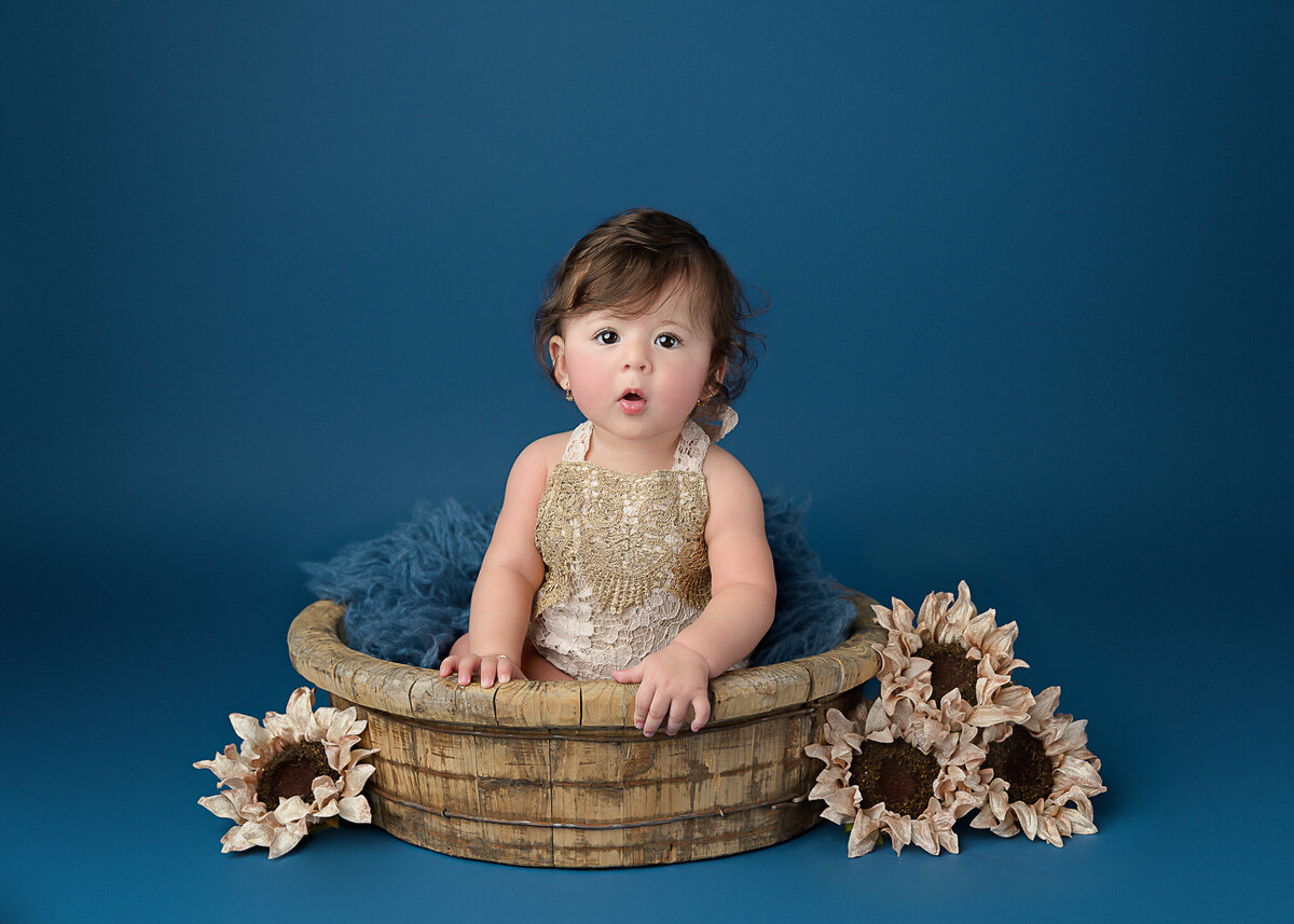 Blue backdrop with sunflowers and wooden bowl for 6 month milestone Little girl in lacy beaded romper