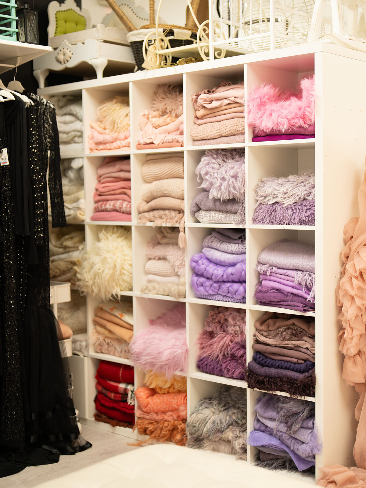 A neatly organized white shelf displays folded pastel and vibrant-colored blankets, creating a cozy and inviting atmosphere. A black sequined dress hangs nearby.