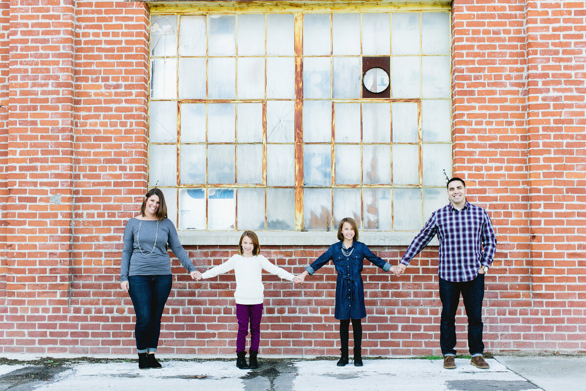 Family of four stand in front of a brick building all holding hands