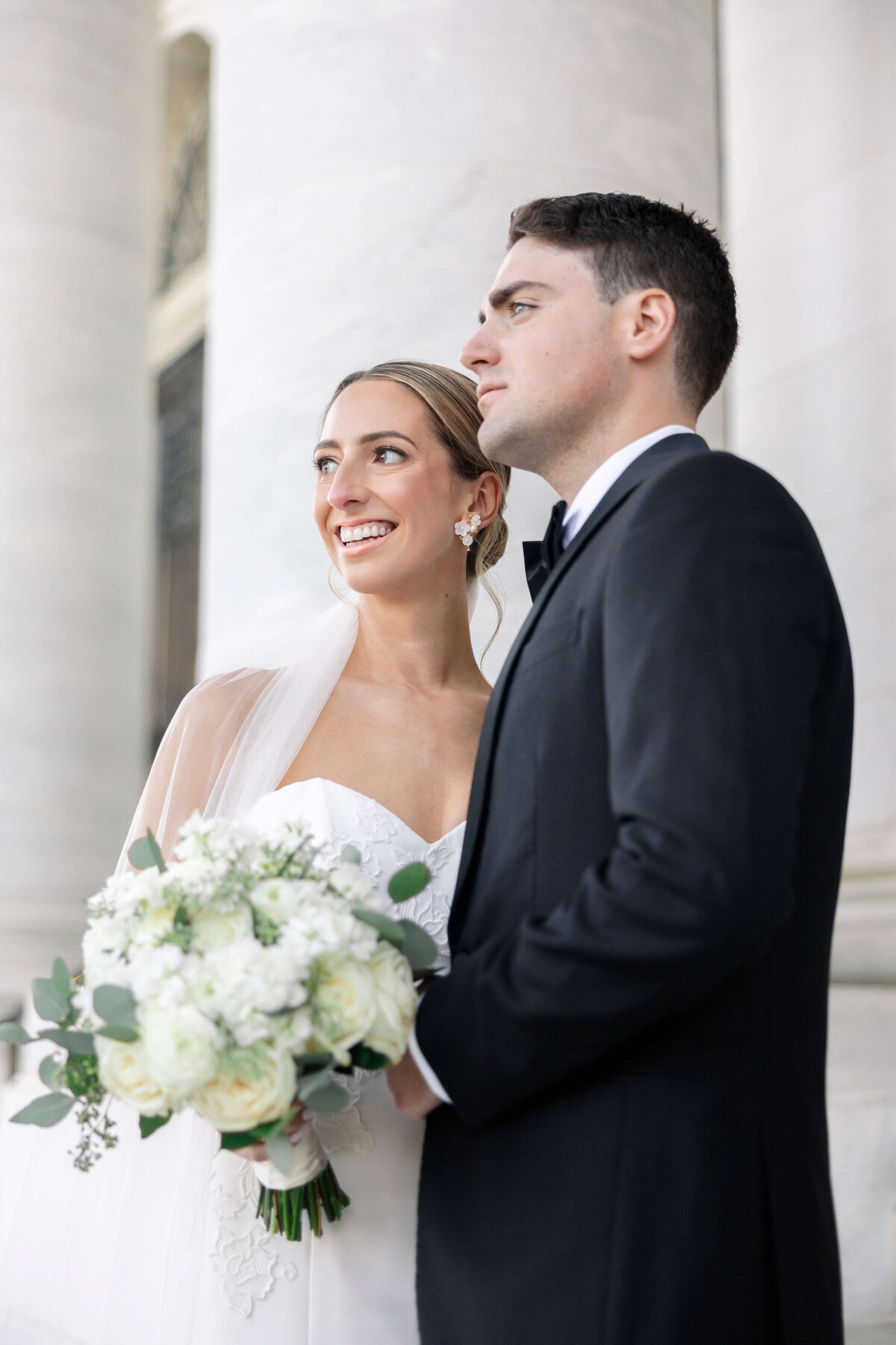 A bride and groom stand in front of a large column. The bride smiles while holding a bouquet of white flowers and greenery, dressed in a white gown and veil. The groom wears a black suit and bow tie, looking forward.