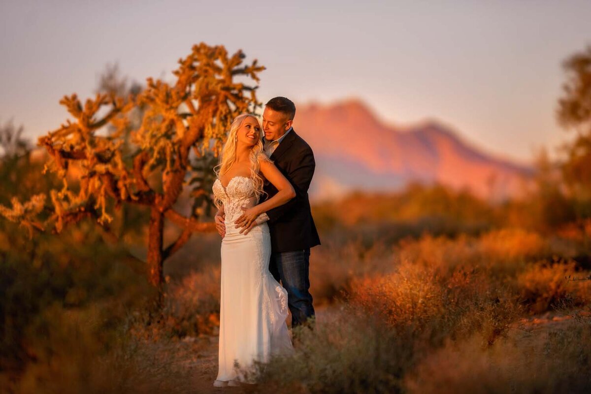 Husband holding his wife during  pink sunset at lost dutchman park surrounded by mountains and cactuses.